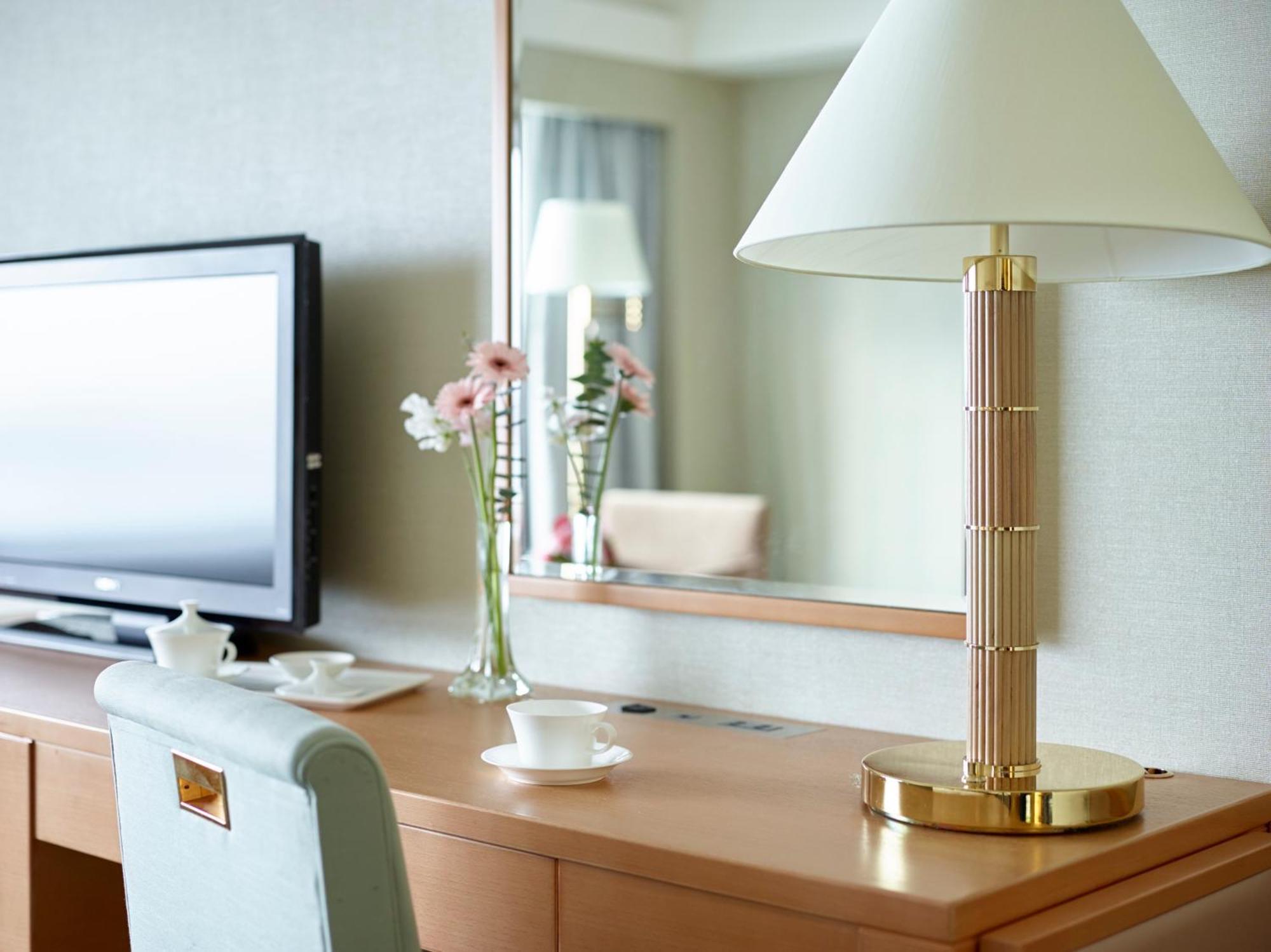 Hotel Okura Kōbe Exterior foto The photo shows an elegantly designed room setup featuring a wooden desk and a large mirror. On the desk, there is a modern lamp with a white shade and a golden base, adding a stylish touch. A television is positioned against the wall on the left sid