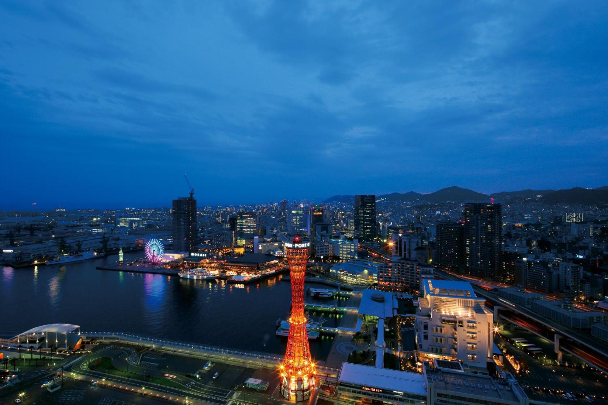 Hotel Okura Kōbe Exterior foto The photo depicts a nighttime cityscape with a view of a harbor. A prominent landmark, likely a tower, is illuminated in bright orange, standing tall against the backdrop of the city. Surrounding buildings and structures are visible, with their light