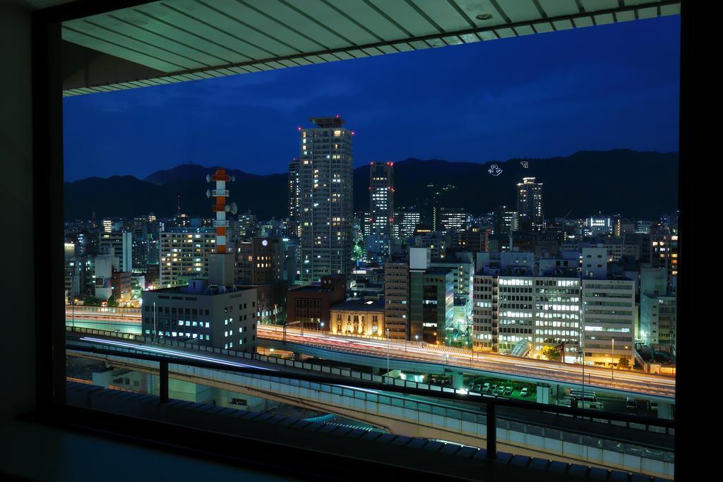 Hotel Okura Kōbe Exterior foto The photo shows a nighttime cityscape viewed from a window. It features a skyline filled with modern buildings, some illuminated with bright lights. In the foreground, there is a balcony railing visible. The view includes a highway with light trails 