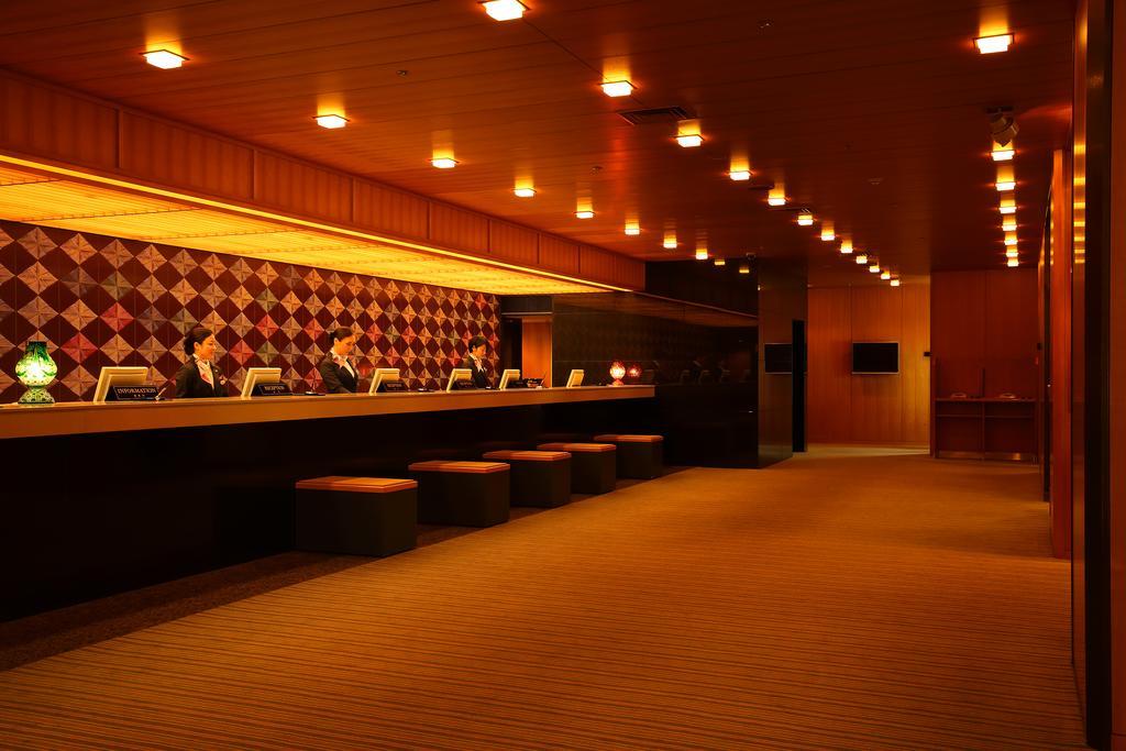 Hotel Okura Kōbe Exterior foto The photo shows the interior of a modern reception area, likely in a hotel or upscale building. There is a long, sleek front desk with several employees dressed in formal attire. Behind the desk is an artistic wall decorated with a diamond pattern in