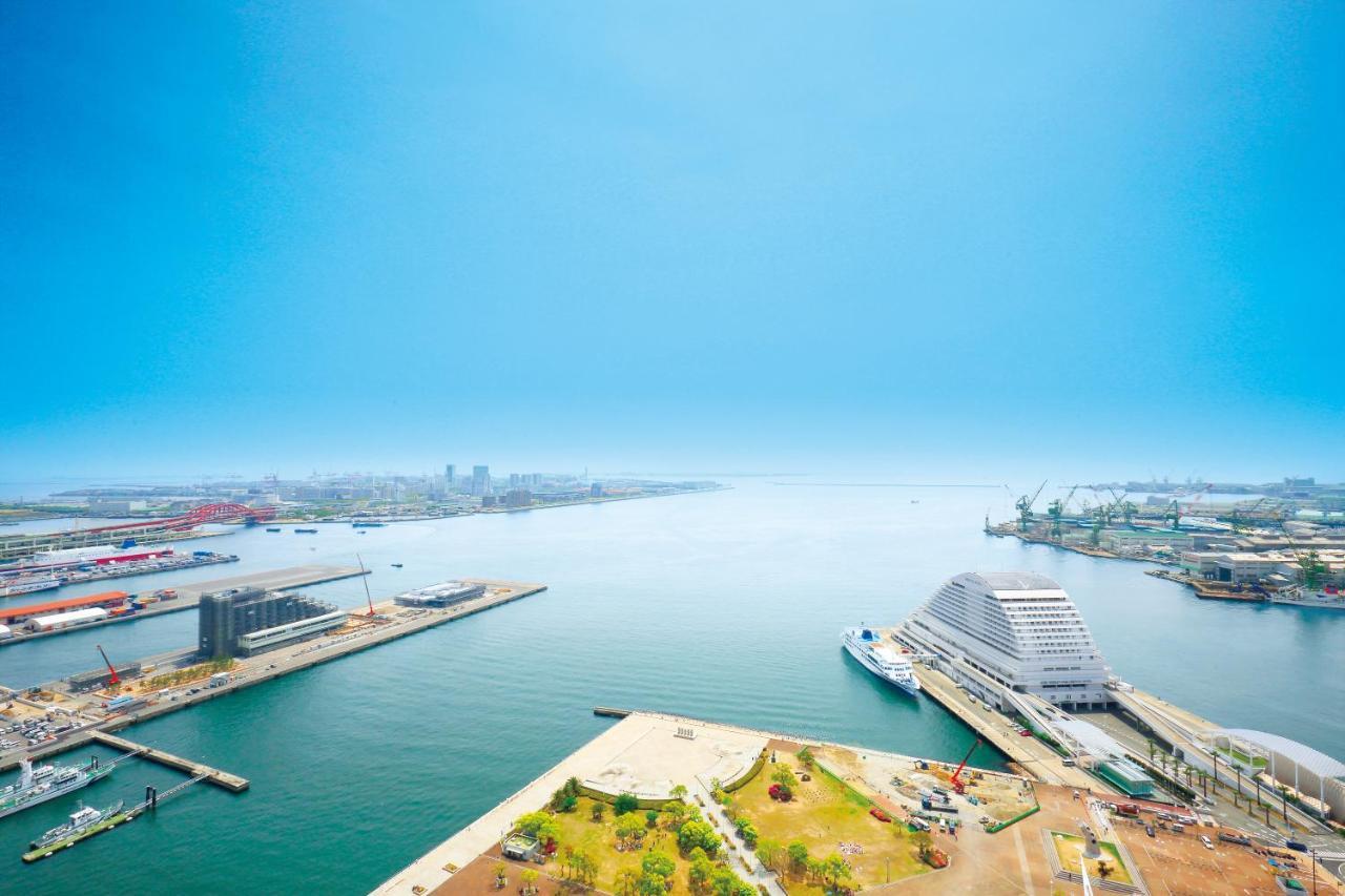 Hotel Okura Kōbe Exterior foto The photo shows a panoramic view of a harbor or waterfront area. In the foreground, there are pathways and green spaces along the shore. The calm water reflects the clear blue sky above, and there’s a prominent building that resembles a modern, curve