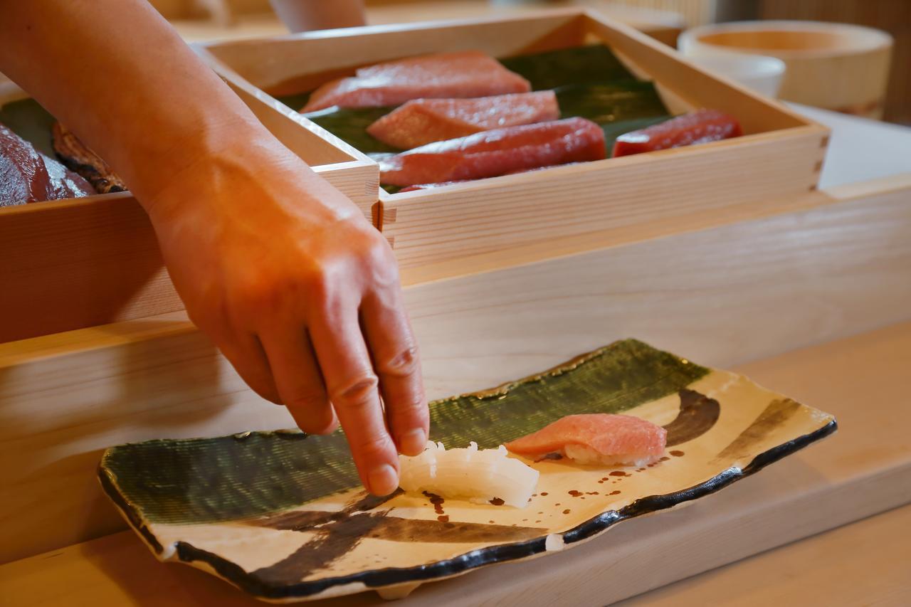 Hotel Okura Kōbe Exterior foto The photo shows a hand carefully placing a piece of sushi onto a decorative plate. The sushi includes a piece of raw fish, likely tuna, and is positioned next to another piece that appears to be rice. In the background, there is a wooden box containi