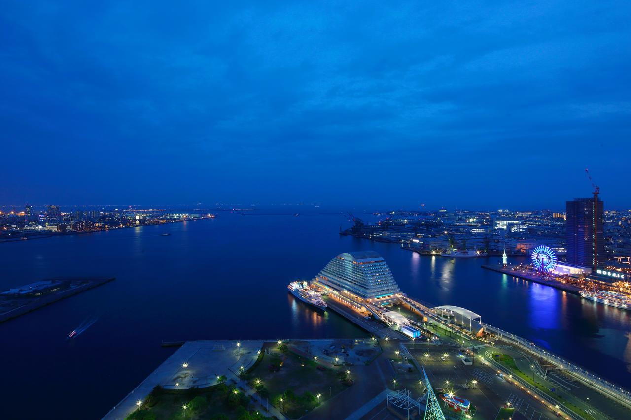 Hotel Okura Kōbe Exterior foto The photo shows a serene evening view of a waterfront cityscape. The scene is illuminated by soft artificial lighting, creating a tranquil ambiance. In the foreground, there are landscaped areas with greenery, and a waterfront promenade lined with li