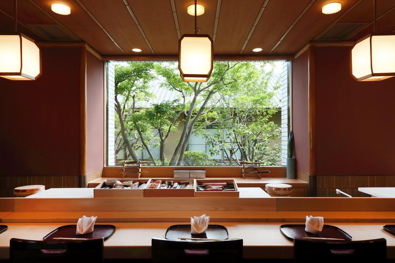 Hotel Okura Kōbe Exterior foto The photo shows a serene dining area, likely in a Japanese restaurant, featuring a wooden counter with several place settings. Light, elegant lanterns hang from the ceiling, creating a warm ambiance. Behind the counter, a large window offers a view o