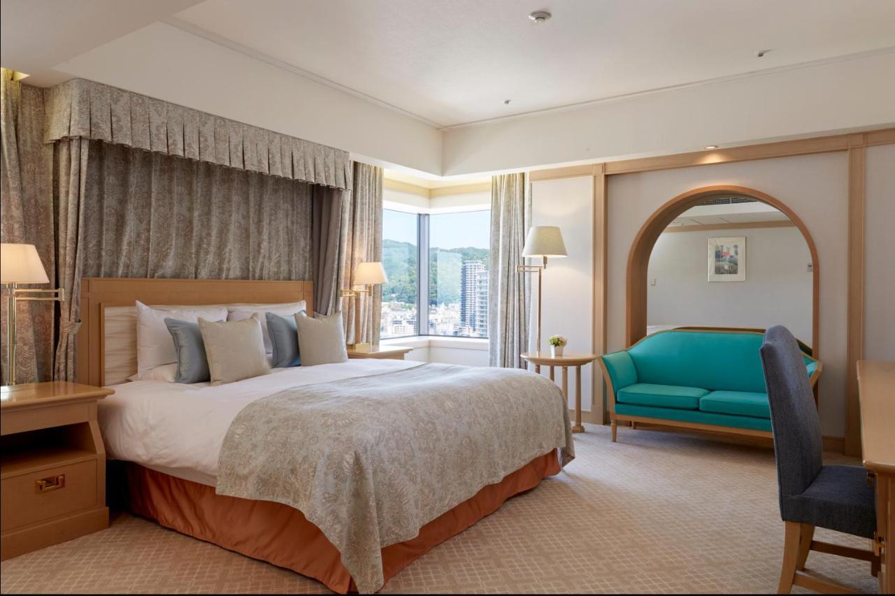 Hotel Okura Kōbe Exterior foto The photo shows a well-appointed hotel room featuring a large bed with decorative pillows and a light-colored bedspread. There is a cozy couch in a teal color placed near a large window, which allows natural light to enter the room. The room has a wa